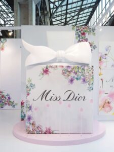 Artistic Miss Dior display featuring a large sculptural perfume bottle with a white bow and floral decorations, designed for luxury perfume branding in a high-end retail setting.