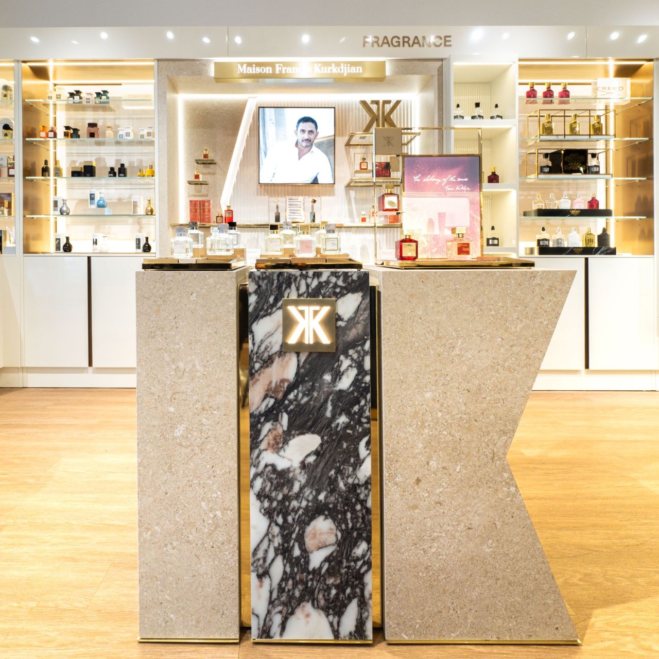 A front-facing view of a luxury custom perfume display counter island featuring a premium marble-veined front panel, a warm-backlit brand logo, and integrated gold metal trim in a high-end department store layout.
