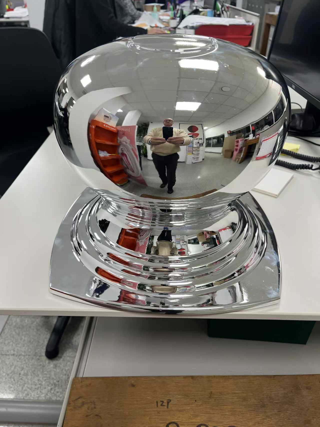 A high gloss chrome finished perfume bottle cap prop sample placed on an office desk showing clear reflection quality and a stepped geometric base.