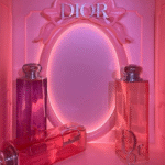 An indoor luxury fragrance pop up retail space featuring large translucent pink perfume bottles and a matching ornate wall mirror frame with glowing backlit lighting.