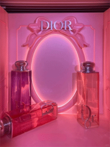 An indoor luxury fragrance pop up retail space featuring large translucent pink perfume bottles and a matching ornate wall mirror frame with glowing backlit lighting.