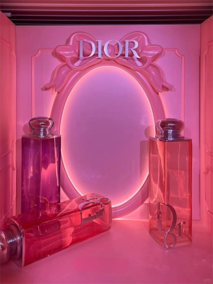 An indoor luxury fragrance pop up retail space featuring large translucent pink perfume bottles and a matching ornate wall mirror frame with glowing backlit lighting.