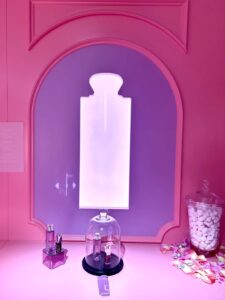 A pink retail product counter with small perfume displays under a glass dome next to a purple arched wall showing a large glowing perfume bottle silhouette.