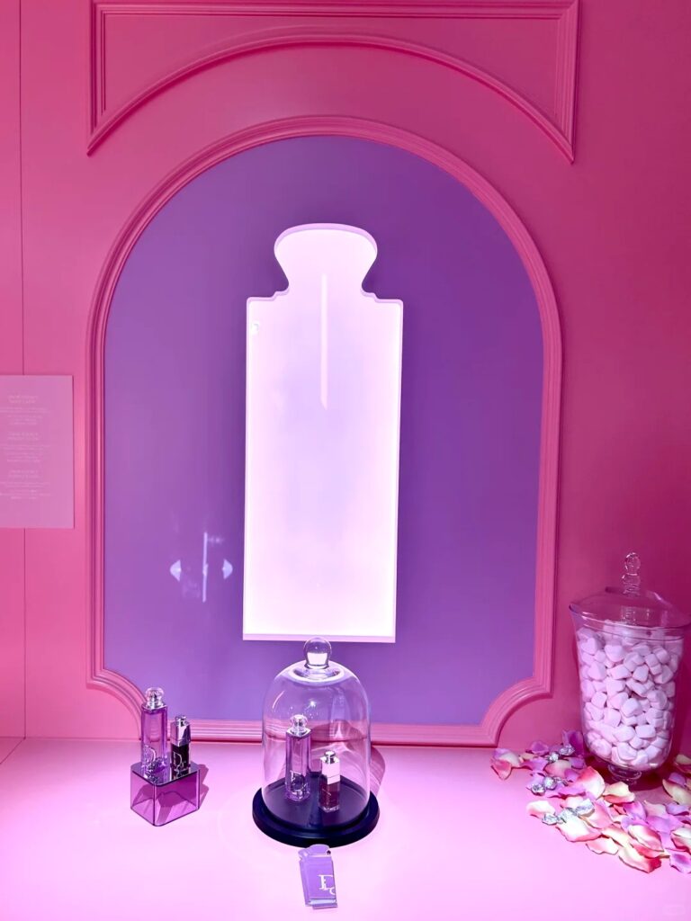 A pink retail product counter with small perfume displays under a glass dome next to a purple arched wall showing a large glowing perfume bottle silhouette.